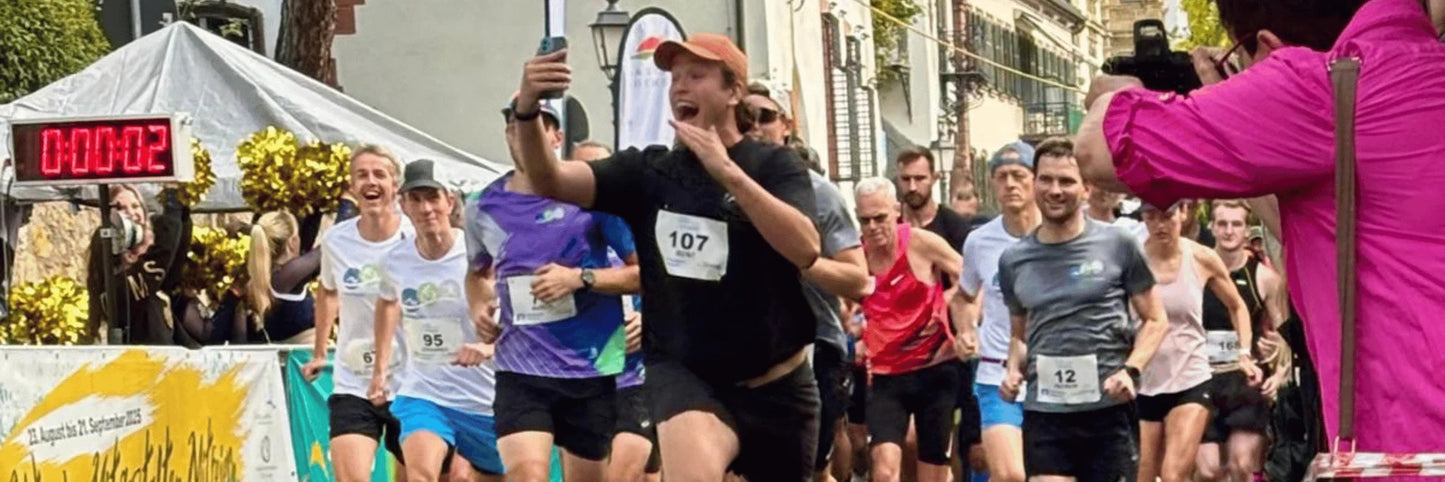 Läufer starten einen Stadtmarathon, einer macht ein Selfie, Zieluhr bei 2 Sekunden, Fotograf im Vordergrund
